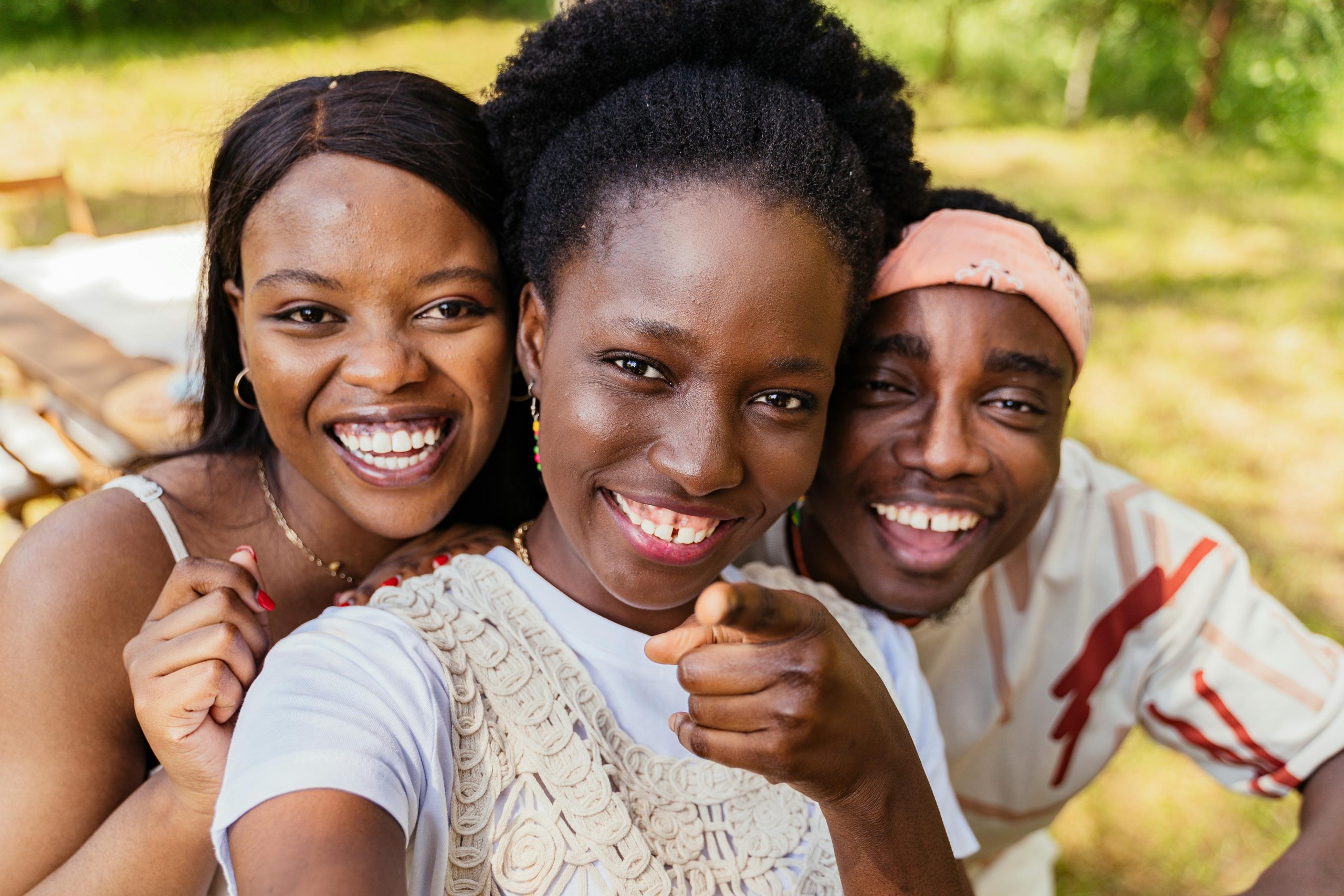 Three friends smiling and pointing at the camera outdoors, enjoying a sunny day.